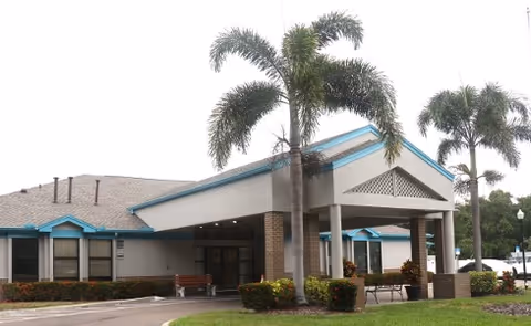 Exterior view of a single-story assisted living facility building with a covered entrance supported by brick columns. There are palm trees and well-maintained landscaping around the building, and a parking area is visible in the background.