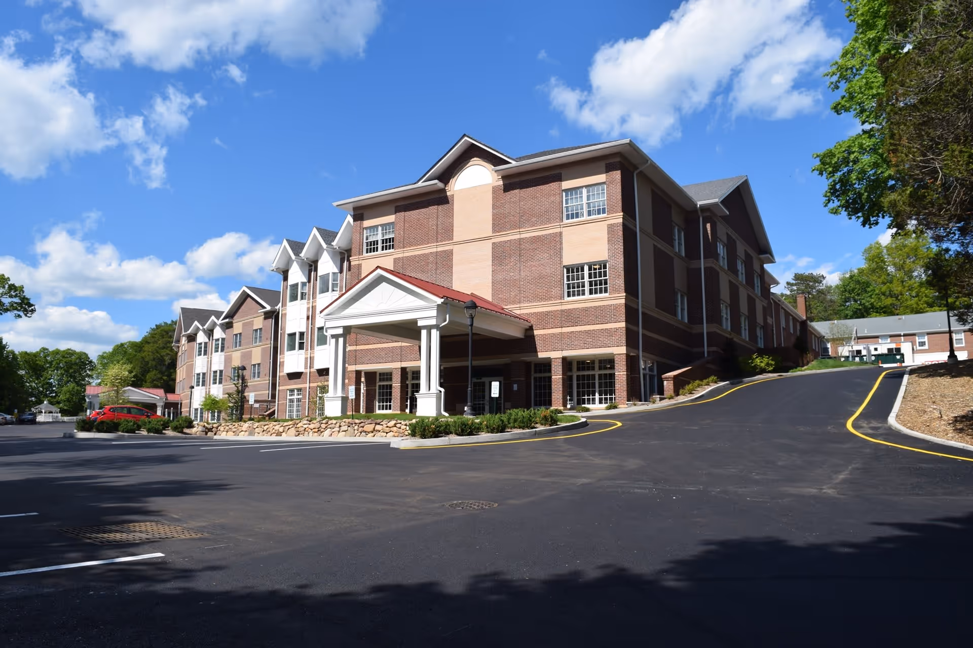Exterior view of a multi-story brick building with white trim and a covered entrance, surrounded by a paved driveway and parking area under a blue sky with scattered clouds.