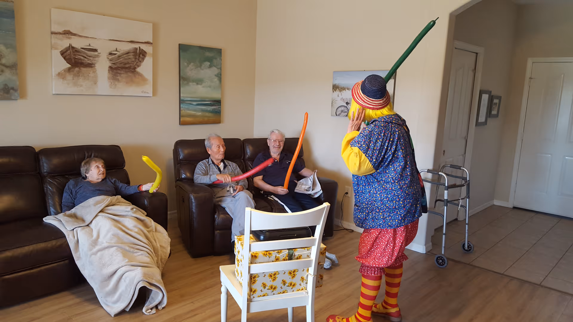 Three elderly people sitting on brown leather chairs in a living room, holding colorful balloon swords, while a person dressed as a clown with a yellow wig, striped socks, and a colorful outfit entertains them. The room has wooden flooring, paintings on the wall, and a walker near the entrance.