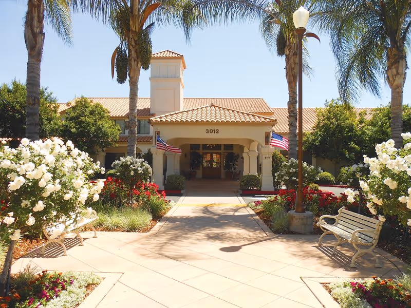 Front exterior view of Oakmont of Escondido Hills facility with a tiled roof entrance, two American flags, palm trees, white flowering bushes, benches, and a paved walkway leading to the door under a clear blue sky.