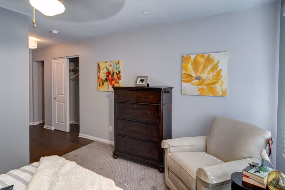 A cozy bedroom corner featuring a beige armchair next to a small round table with books and a decorative bird figurine. A dark wooden chest of drawers stands against the wall, flanked by two colorful floral paintings. The room has light gray walls and a carpeted floor, with an open closet door visible in the background.