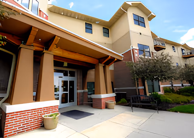 Entrance of a multi-story senior living facility with a covered porch supported by large columns, a glass door, potted plants, a bench, and landscaped greenery under a partly cloudy sky.