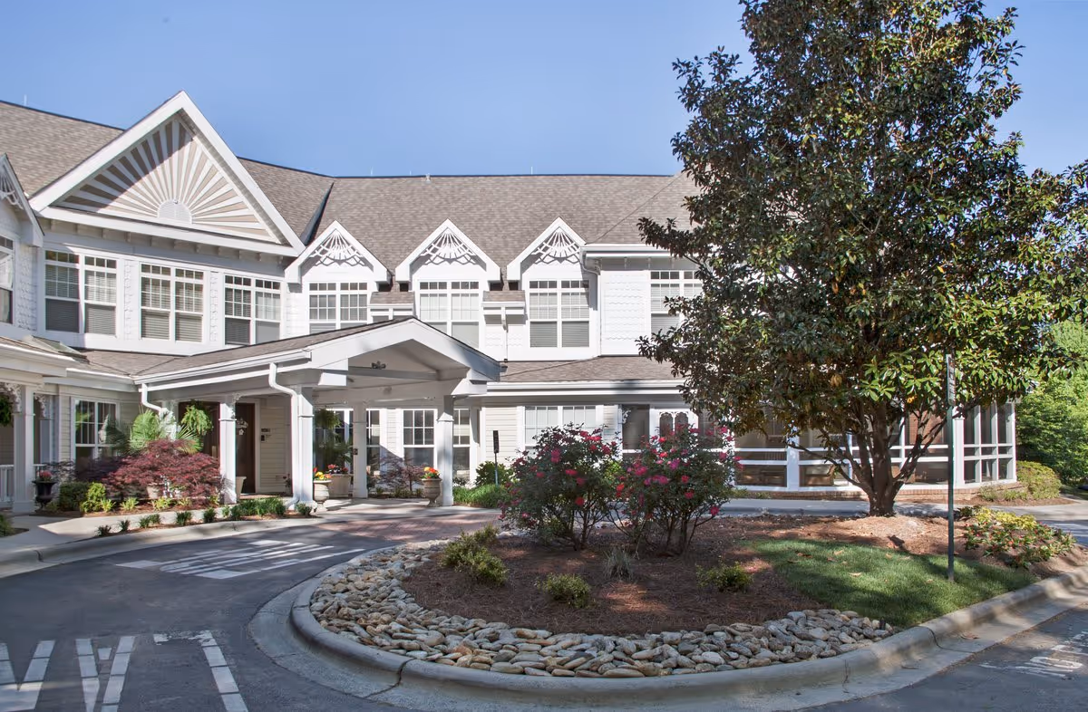 Front entrance of a two-story senior living building with a covered porte-cochère, landscaped circular driveway and trees.