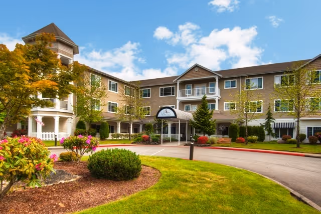 Exterior view of a three-story senior living facility with beige siding, white trim, and a covered entrance. The building is surrounded by well-maintained landscaping including green grass, bushes, and flowering plants under a partly cloudy blue sky.