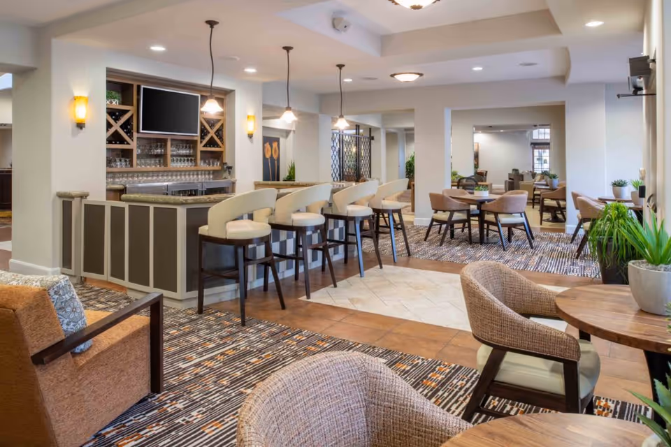 A spacious and well-lit common area in a senior living facility featuring a bar with high chairs, multiple round tables with cushioned chairs, potted plants, and modern lighting fixtures. The room has a mix of carpet and tile flooring and a neutral color palette.