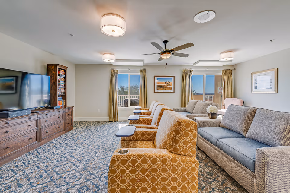 Bright furnished common living room with rows of upholstered armchairs and sofas facing a large TV, windows, and a ceiling fan.