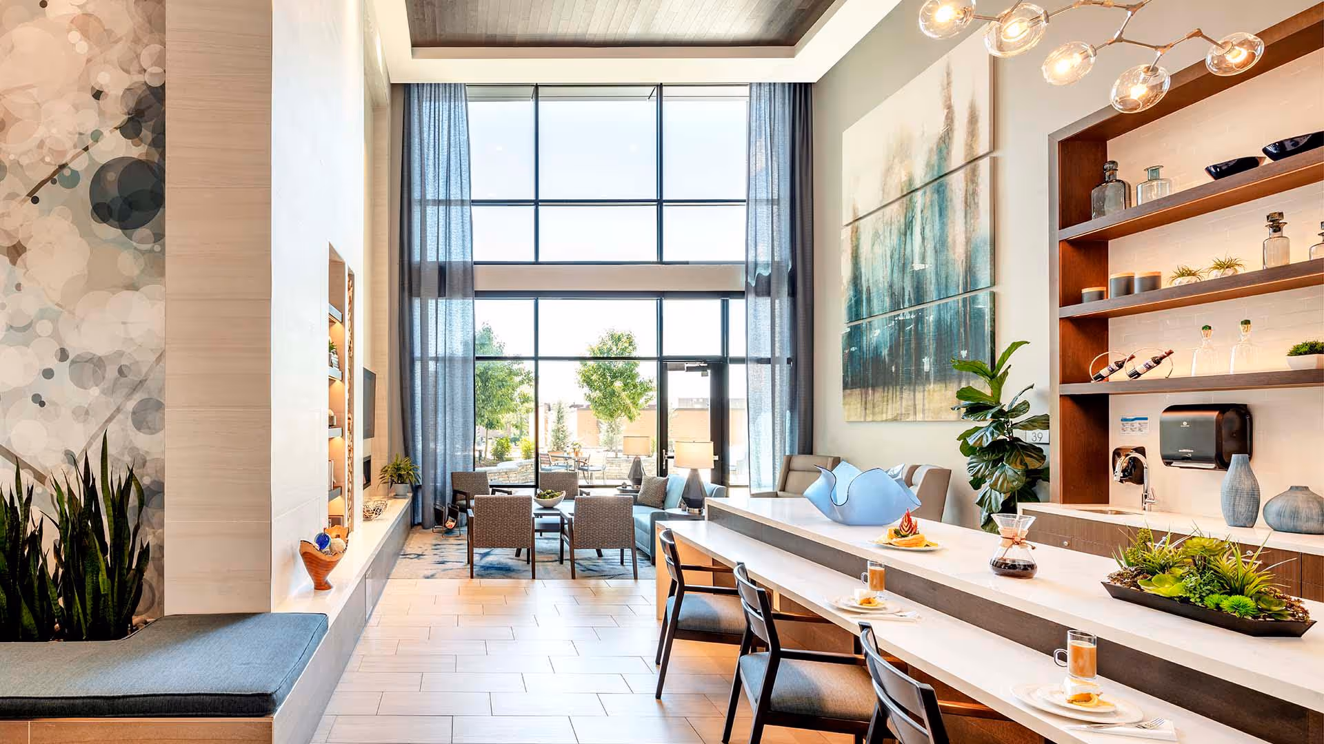 Bright modern senior living common area with tall windows, seating groups, and a long counter with stools.