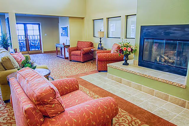 A cozy senior living facility common area with patterned red armchairs and a sofa arranged around a tiled fireplace. The room features a carpet with a floral design, a side table with a lamp, a vase of flowers on the fireplace ledge, and large windows and doors letting in natural light.