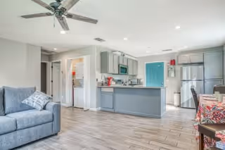 Bright and modern open-concept living space featuring a gray sofa with patterned pillows, a ceiling fan, a kitchen with gray cabinets, stainless steel appliances, and a blue door in the background. A dining table with colorful chairs is partially visible on the right.