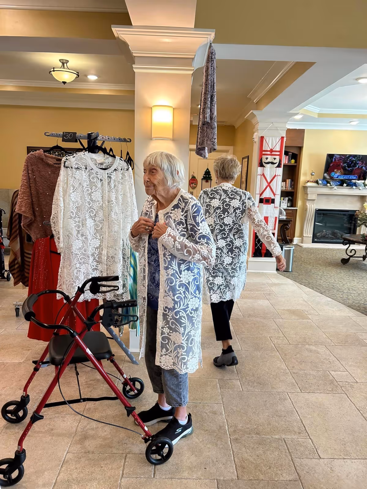 An elderly woman with white hair stands indoors near a clothing rack, trying on a white lace cardigan. She uses a red walker for support. The room has beige tiled floors, yellow walls, and a fireplace with a TV mounted above it. Another elderly woman is seen walking away in the background, also wearing a similar white lace cardigan.