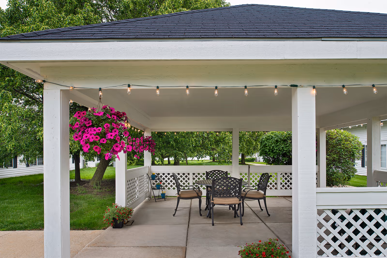 Covered outdoor patio area with a table and four chairs, hanging baskets of vibrant pink flowers, string lights hanging from the ceiling, and green trees and bushes surrounding the space.