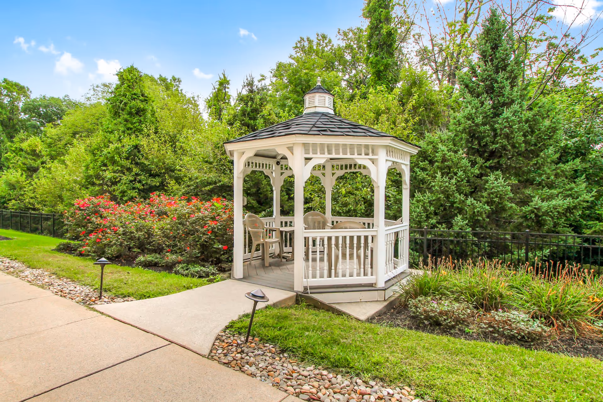 A white wooden gazebo with a dark shingled roof situated in a garden area surrounded by green trees, shrubs, and flowering plants. There are several plastic chairs inside the gazebo. A concrete pathway with small garden lights leads to the gazebo.
