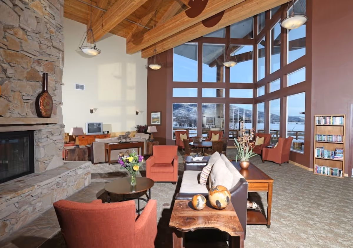 Spacious living room with large floor-to-ceiling windows offering a view of snow-covered mountains. The room features a stone fireplace on the left, wooden ceiling beams, and warm lighting fixtures. There are several comfortable seating options including red armchairs and a beige sofa with decorative pillows. A wooden coffee table with a vase of flowers and a side table with decorative spheres are also visible. A bookshelf filled with books is positioned against the right wall.