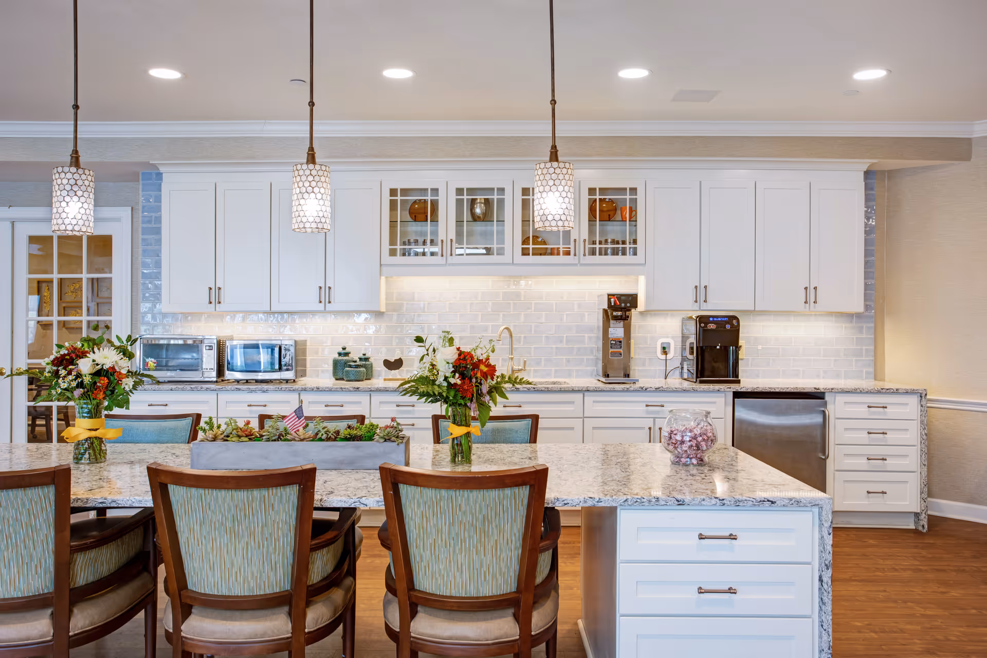 Bright communal kitchen with a large granite island, pendant lights, white cabinets, appliances, and chairs with floral centerpieces.