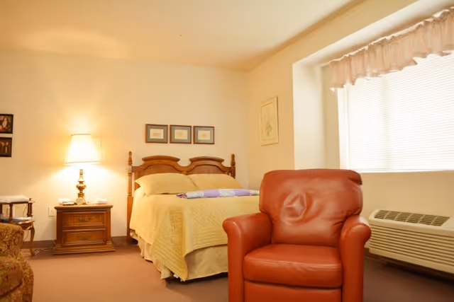 A cozy bedroom with a wooden bed frame, a yellow bedspread, and a folded quilt on top. There is a red leather armchair in the foreground, a wooden nightstand with a lamp and some items on it, and a window with blinds and a valance. The walls are decorated with framed pictures.