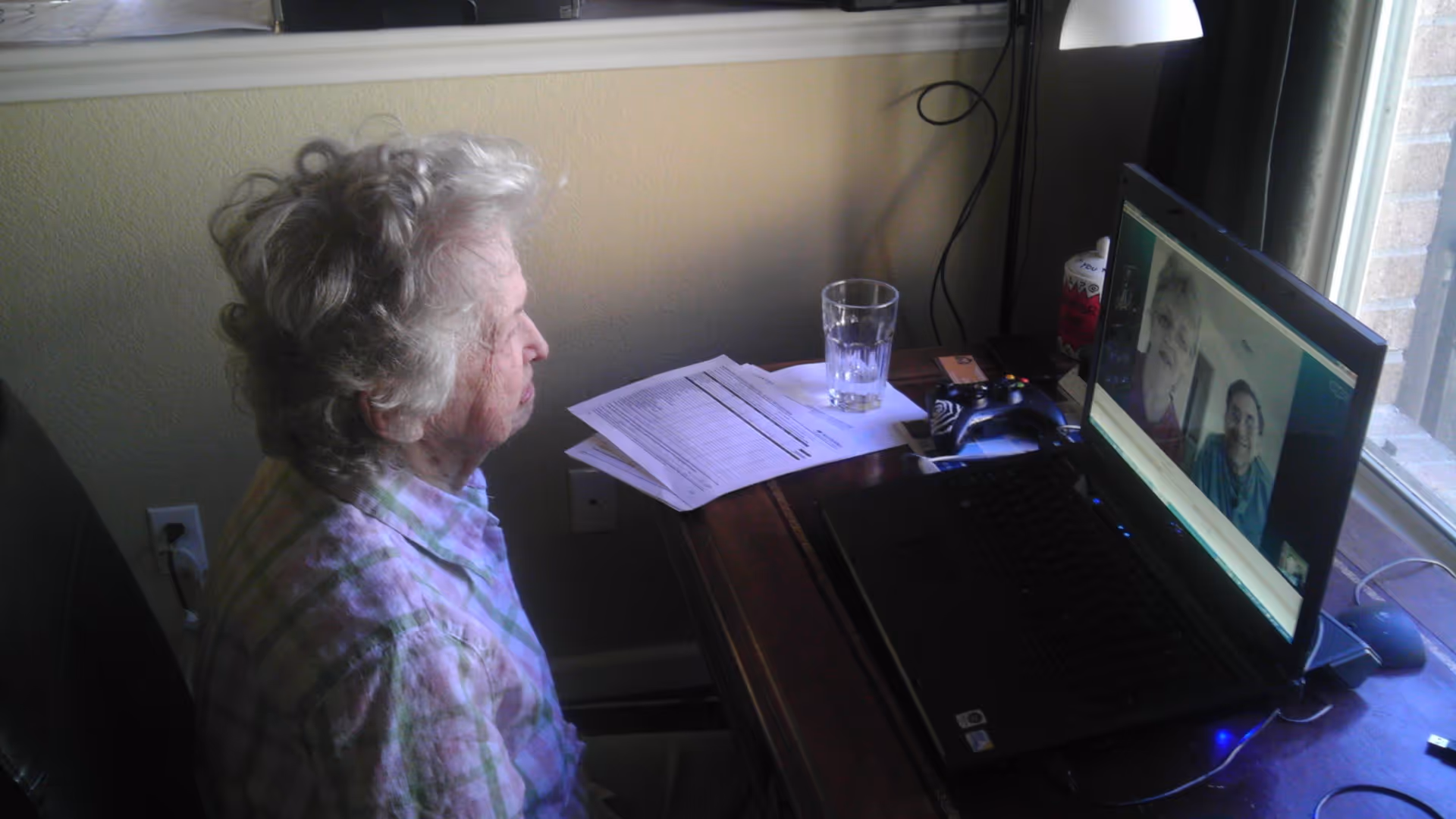 An elderly woman with curly gray hair sits at a wooden desk in front of a laptop, engaged in a video call with two other elderly individuals visible on the screen. The desk has papers, a glass of water, a game controller, and a lamp. The room has a window with curtains and a light-colored wall.