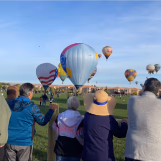 A group of people standing behind a wooden fence watching colorful hot air balloons being inflated and floating in the sky over a grassy field with houses in the background.