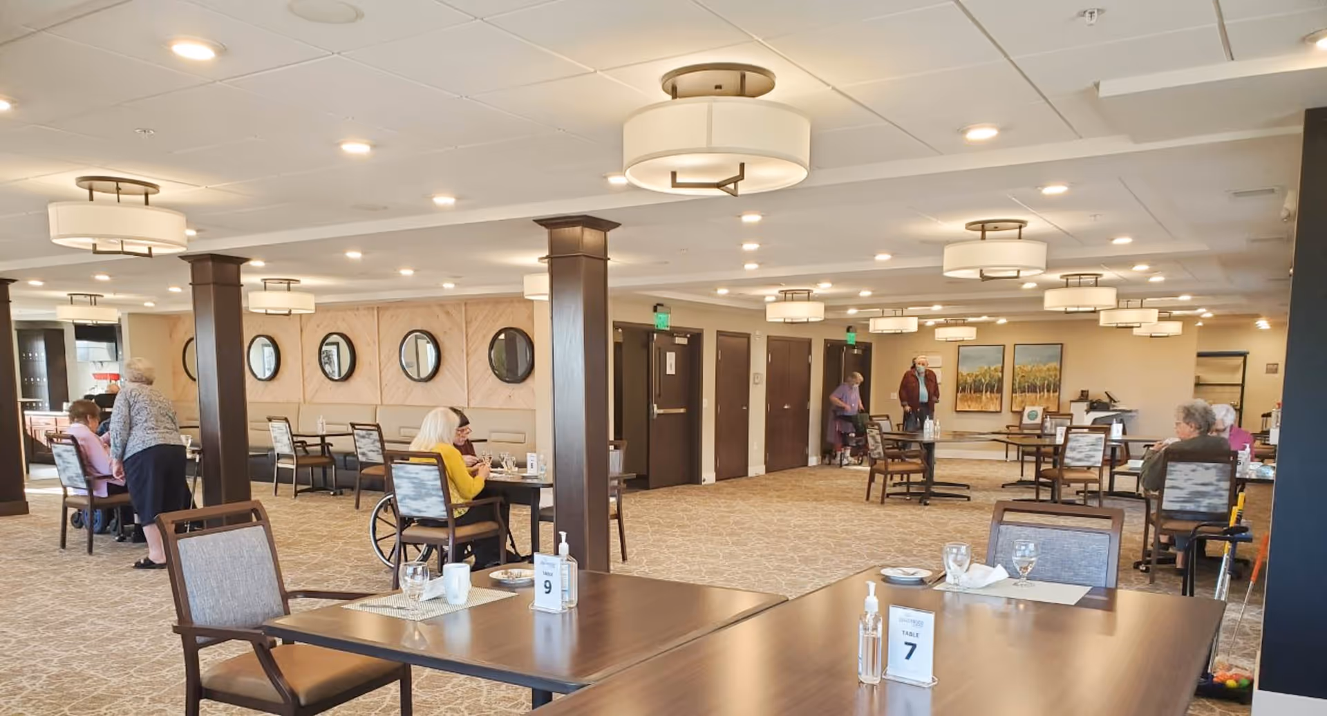 Spacious dining room with multiple tables and chairs, overhead drum lights, and several elderly residents dining and socializing.