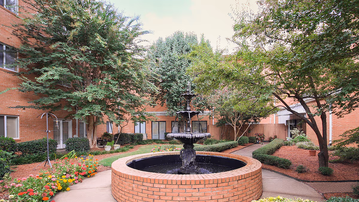 A peaceful outdoor courtyard at Holiday Chateau Ridgeland featuring a central brick fountain surrounded by colorful flowers, green shrubs, and trees. The courtyard is enclosed by a brick building with windows and doors visible.