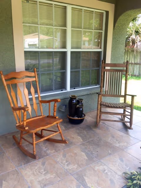 Two wooden rocking chairs on a tiled porch in front of a window with white blinds, next to a small decorative water fountain with three black ceramic pots.
