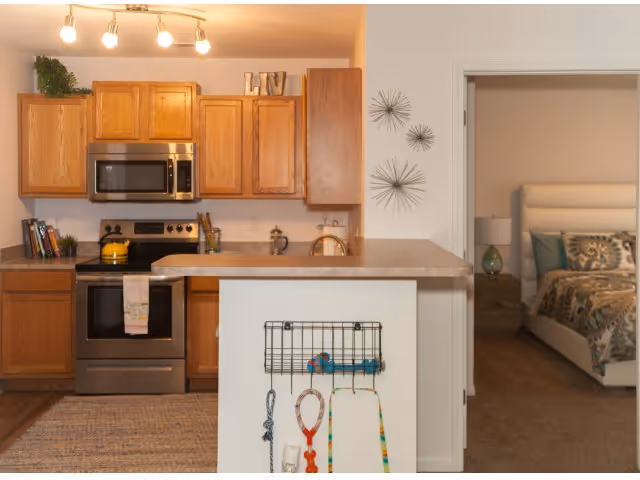 Interior view of a kitchen with wooden cabinets, a stainless steel microwave and oven, a countertop with a small overhang, and a wall-mounted organizer holding various items. To the right, an open doorway leads to a bedroom with a bed, nightstand, lamp, and decorative pillows.