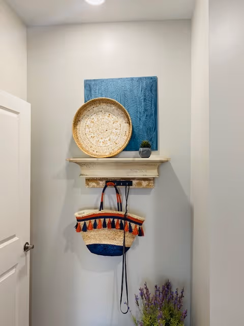 A small wall area with a white door on the left, featuring a rustic wooden shelf mounted on the wall. On the shelf, there is a round woven basket and a small potted plant. Below the shelf, a colorful woven bag with tassels is hanging. At the bottom right, there is a bunch of purple flowers in a container.