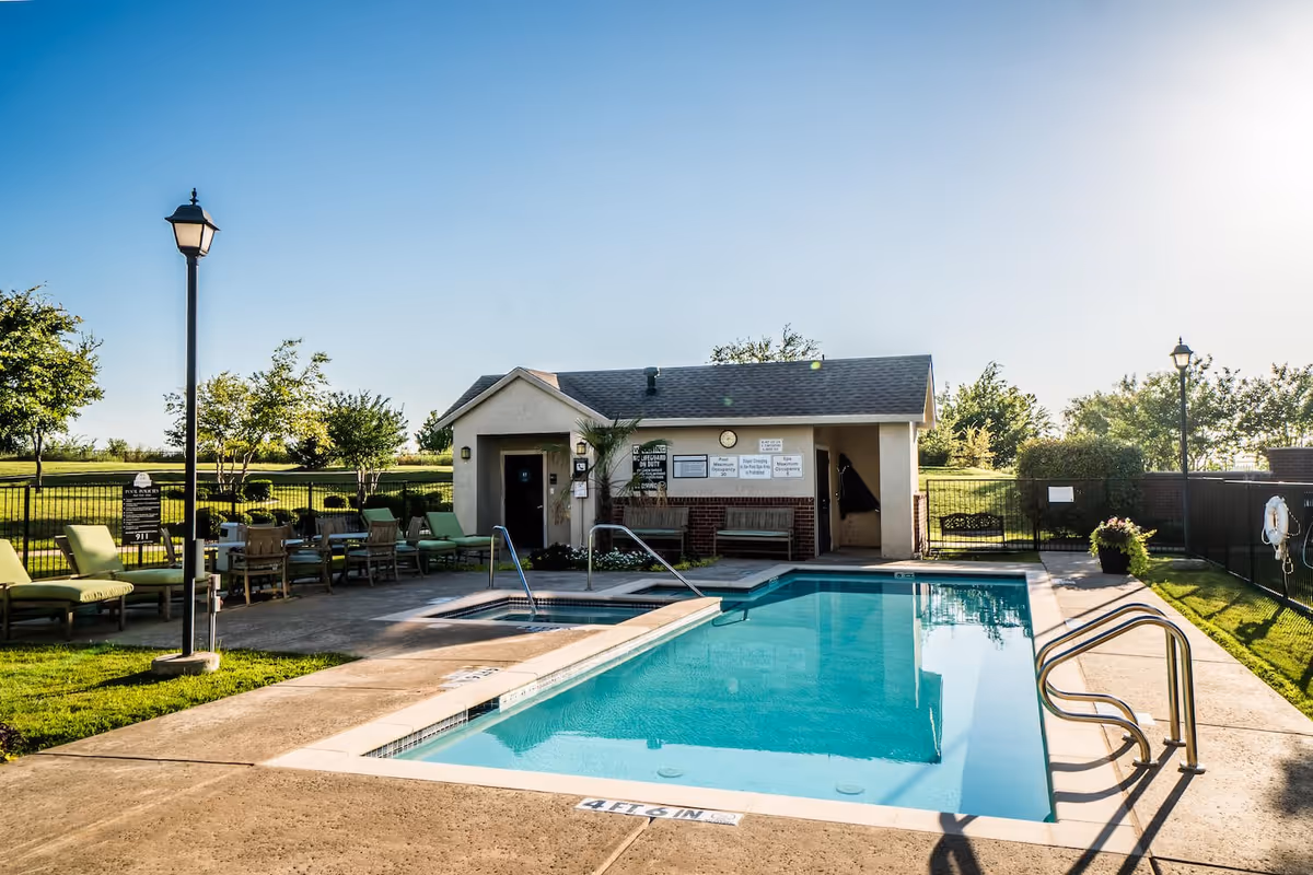 Outdoor swimming pool area with clear blue water, surrounded by a concrete deck with lounge chairs and tables. A small building with signs and benches is visible in the background, along with green grass, trees, and a clear blue sky.