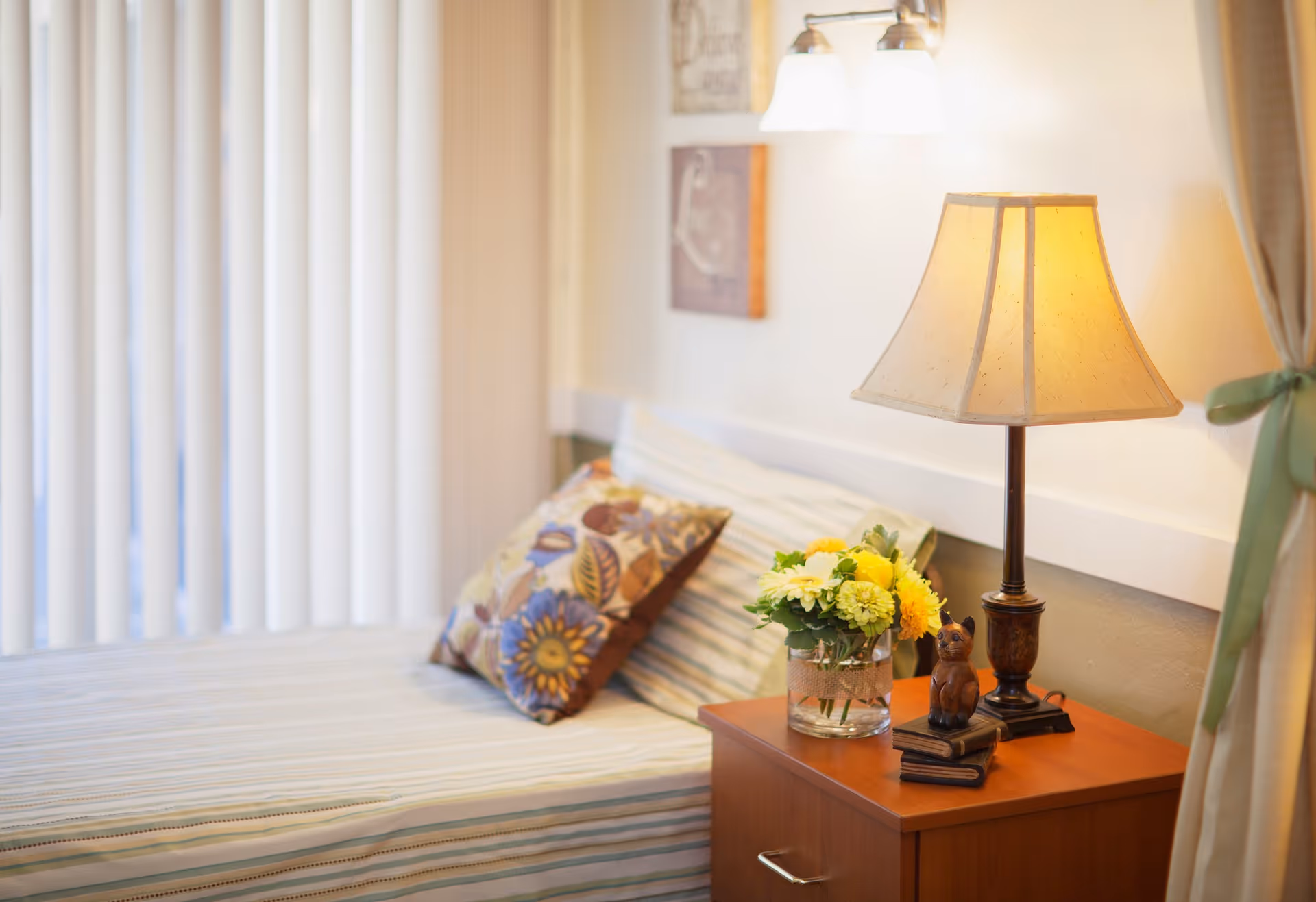 A cozy bedroom corner with a bed featuring striped bedding and decorative pillows. Next to the bed is a wooden nightstand holding a table lamp, a small figurine of a cat, and a glass vase with yellow and white flowers. Vertical blinds cover the window, and light-colored curtains are tied back with a green ribbon.