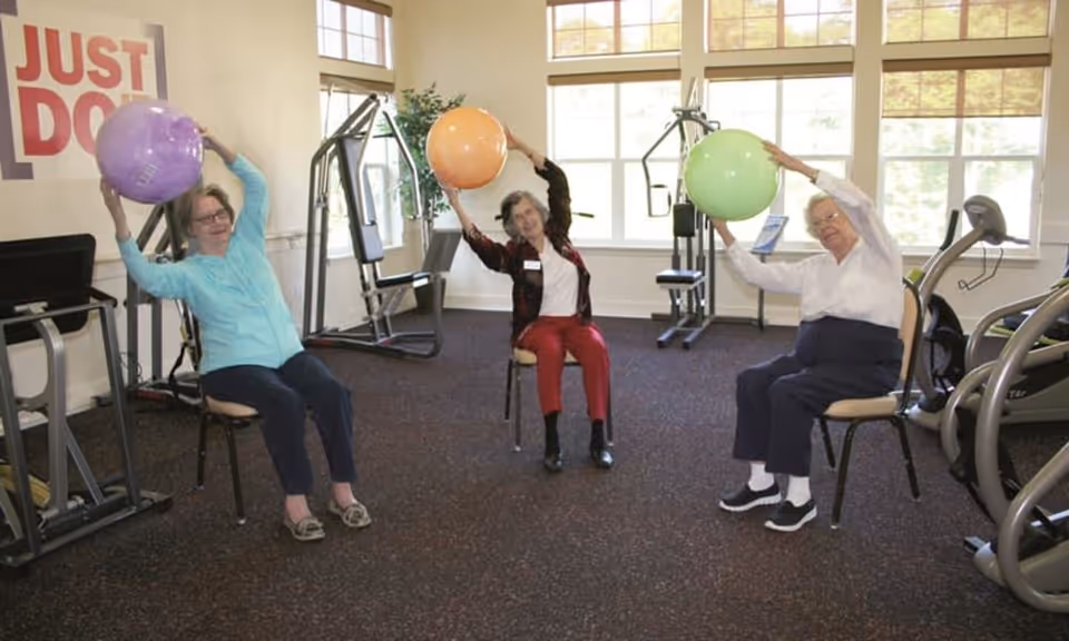 Three elderly women seated on chairs in a fitness room holding large exercise balls above their heads, with exercise equipment and large windows in the background. A sign on the wall reads 'JUST DO IT'.