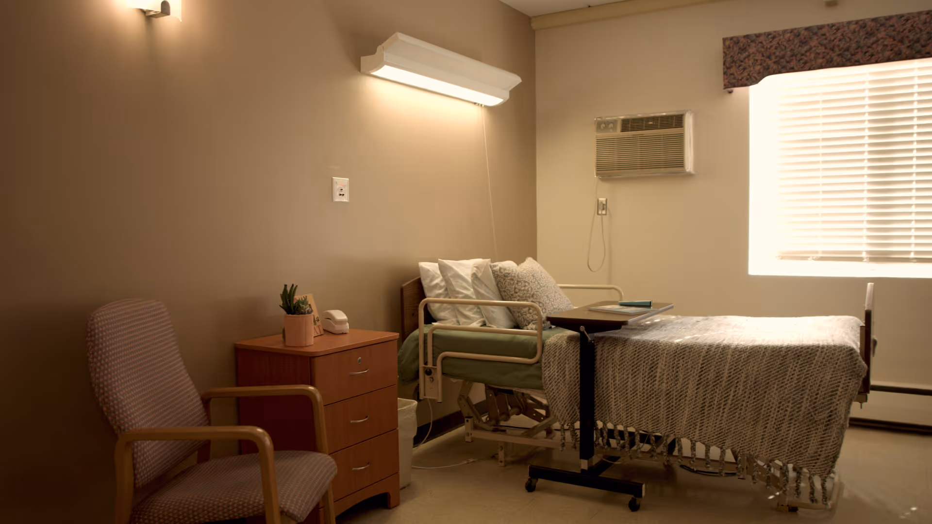 A simple, well-lit bedroom in a senior living facility featuring a hospital-style adjustable bed with pillows and a blanket, a wooden nightstand with a small plant and a telephone, a cushioned armchair, a window with blinds and a valance, and a wall-mounted air conditioning unit.