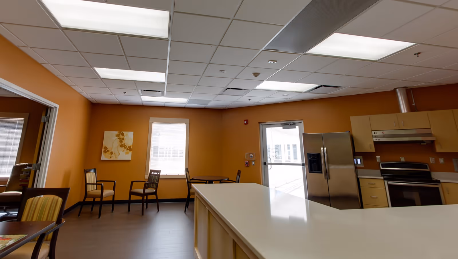 Interior view of a kitchen and dining area with orange walls, a white countertop island, stainless steel refrigerator and oven, several chairs and tables, and a window letting in natural light.