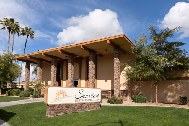 Exterior view of Sunview Health and Rehabilitation Center building with stone pillars and a sign displaying the facility name on a well-maintained lawn under a partly cloudy sky.