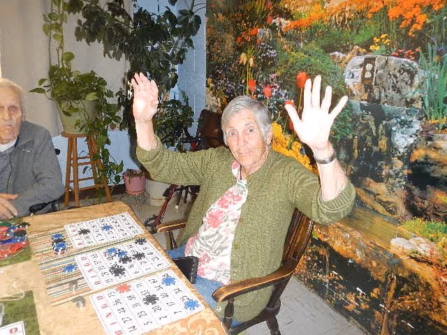 Two elderly women sitting at a table playing bingo. One woman is raising both hands and smiling, while the other woman sits quietly. The room has a large colorful mural of a garden with flowers and rocks on the wall behind them, and there are some potted plants in the background.
