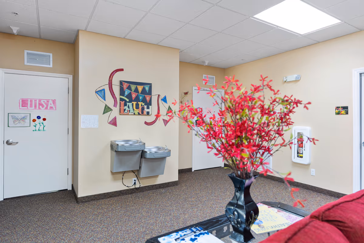 Interior view of a common area in an assisted living facility with beige walls, a double water fountain mounted on the wall, colorful wall decorations including a sign that says 'Live Love Laugh', a door with the name 'Luisa' and children's artwork, a fire extinguisher cabinet, and a vase with red flowers on a table in the foreground.