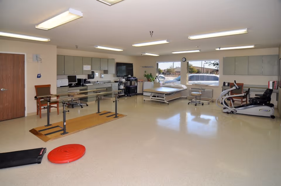 A spacious rehabilitation or physical therapy room with parallel bars for walking exercises, a treatment bed, exercise equipment including a recumbent bike, chairs, cabinets, and large windows letting in natural light.