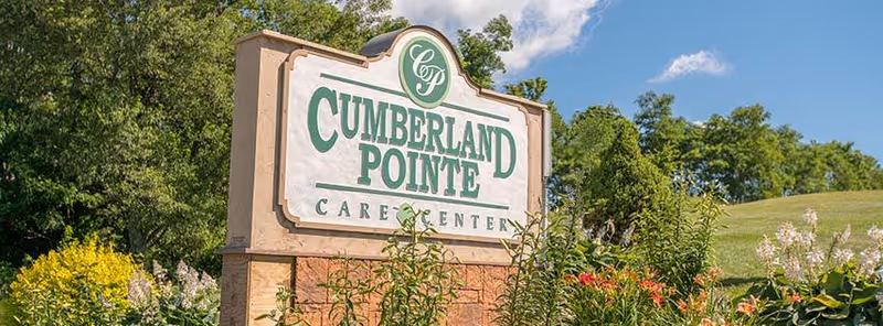 Stone sign for Cumberland Pointe Care Center surrounded by green plants and flowers with trees and a blue sky in the background.