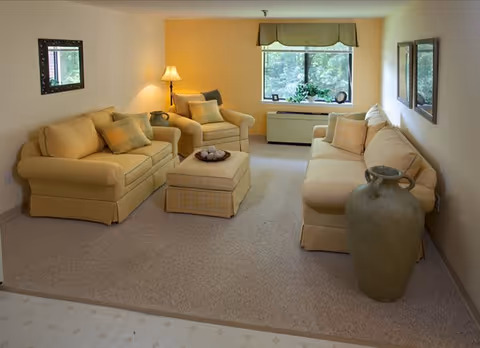 Sunny living room with matching yellow sofa and armchairs arranged around an ottoman, a window with a valance, lamp, and large decorative vase.