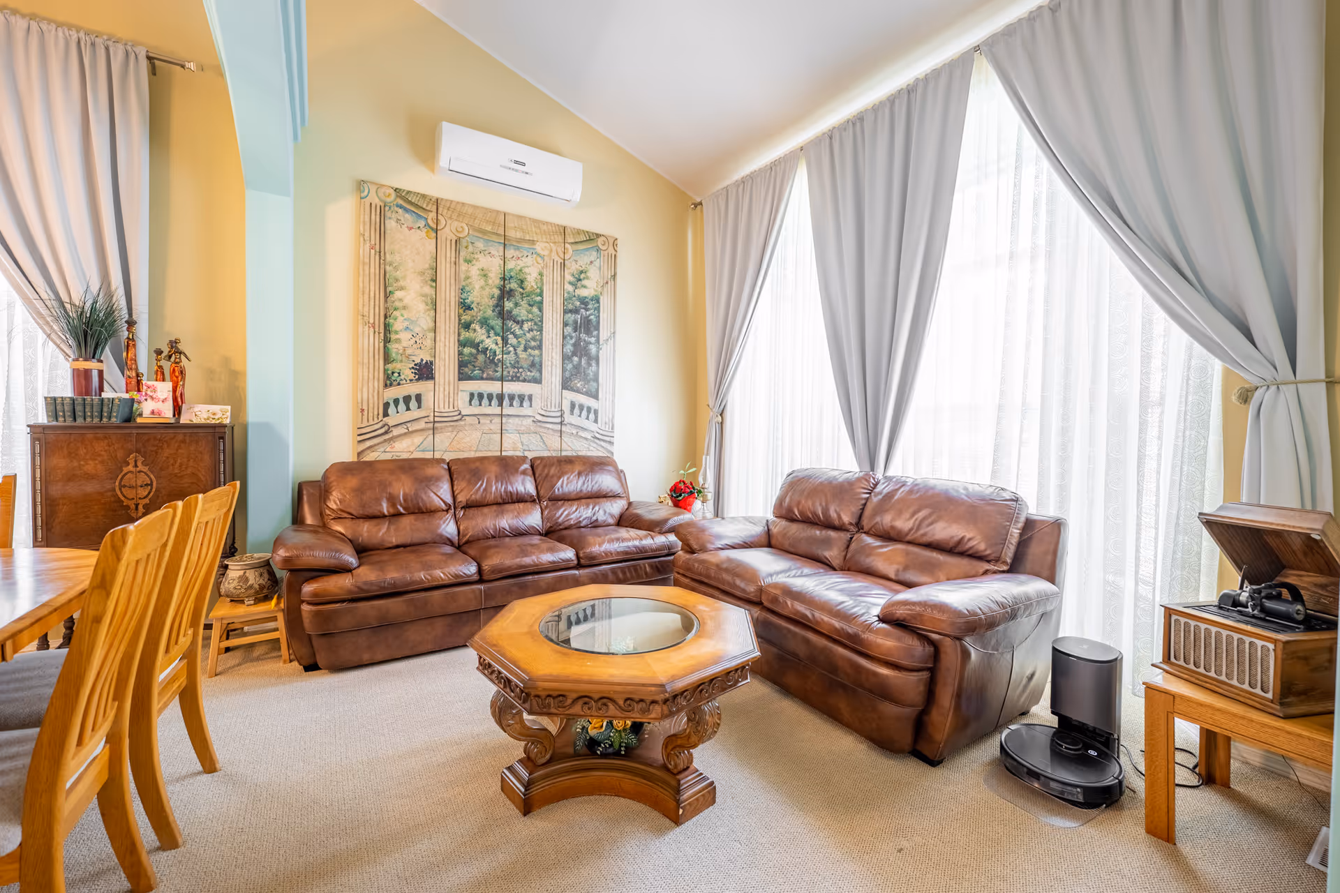 A cozy living room with two brown leather sofas arranged around a wooden octagonal coffee table with a glass top. Large windows with sheer white curtains and gray drapes allow natural light to fill the room. A decorative wall panel depicting a scenic view is mounted above one sofa. To the right, there is a wooden side table with a vintage record player and a small black robot vacuum cleaner on the floor. On the left, a wooden dining table with chairs and a cabinet with decorative items are visible.