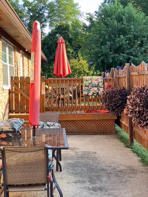 Outdoor patio area with a glass-top table and chairs featuring floral cushions, a closed red umbrella on the table, a wooden deck with a red umbrella, and a wooden fence surrounding the space with green trees in the background.