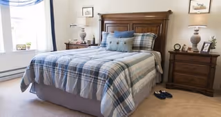 A bedroom with a large wooden bed frame and a bed made with a blue and gray plaid comforter and multiple pillows. There are two wooden nightstands on either side of the bed, each with a lamp and decorative items. A window with sheer white curtains and a blue valance is visible on the left side of the room. A pair of slippers is placed on the carpeted floor near the bed.