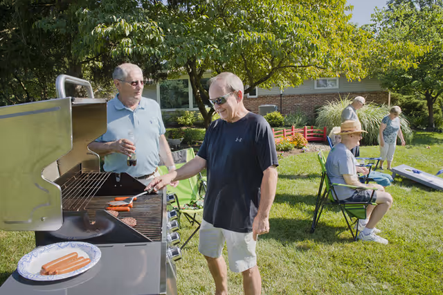 A group of elderly people enjoying an outdoor barbecue in a garden area. One man is grilling hot dogs on a large gas grill while another man stands beside him holding a drink. In the background, three other seniors are seated and standing near a lawn game setup, surrounded by green grass, trees, and a building.