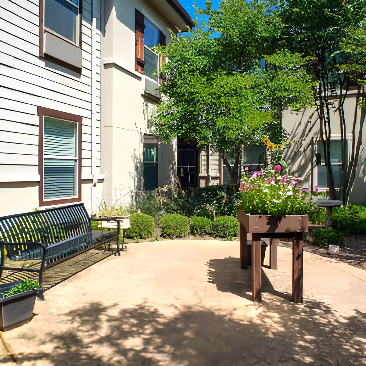 Outdoor courtyard area with a black metal bench on the left, a raised garden bed with blooming flowers in the center, surrounded by green bushes and trees, and beige building walls with windows in the background under a clear blue sky.