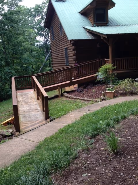 A wooden cabin-style building with a green metal roof surrounded by trees and greenery. There is a wooden ramp with railings leading up to the entrance, and a concrete walkway curves through a garden area with plants and mulch.