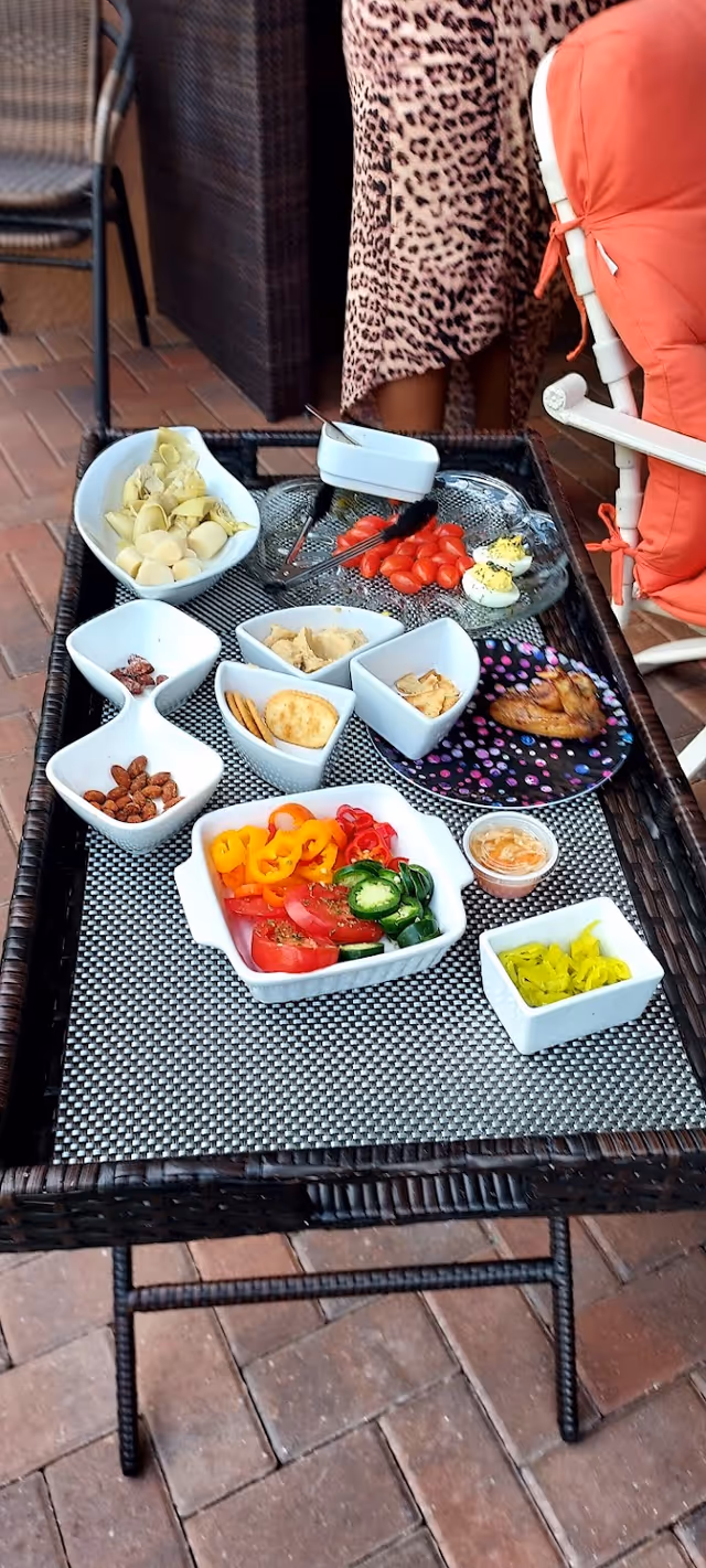 A wicker table with a variety of small dishes containing snacks and appetizers including sliced tomatoes, bell peppers, jalapenos, crackers, nuts, deviled eggs, and a small dish of dip. The table is set on a brick patio with a person wearing a leopard print skirt and an orange cushioned chair partially visible in the background.