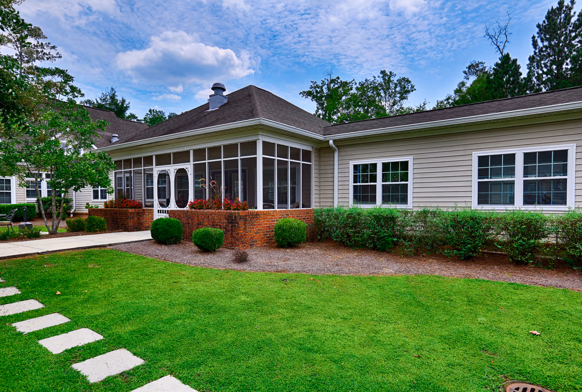 Exterior view of a single-story building with beige siding and a dark shingled roof, featuring a screened porch with white framing and a brick base. The building is surrounded by neatly trimmed bushes, a green lawn, and a stone pathway leading to the entrance. Trees and a partly cloudy blue sky are visible in the background.