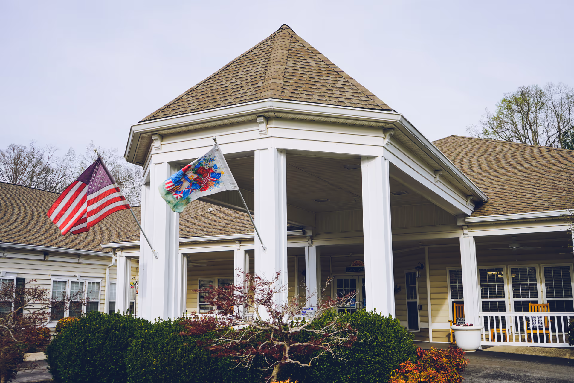 Front entrance of a senior living facility with white columns, an American flag and a decorative flag flying over a covered porch.