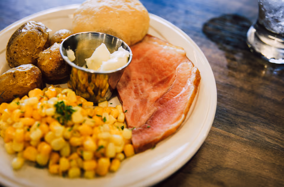 A plate of food with a slice of ham, corn, roasted potatoes, a bread roll, and a small metal cup of butter on a wooden table next to a glass of water.