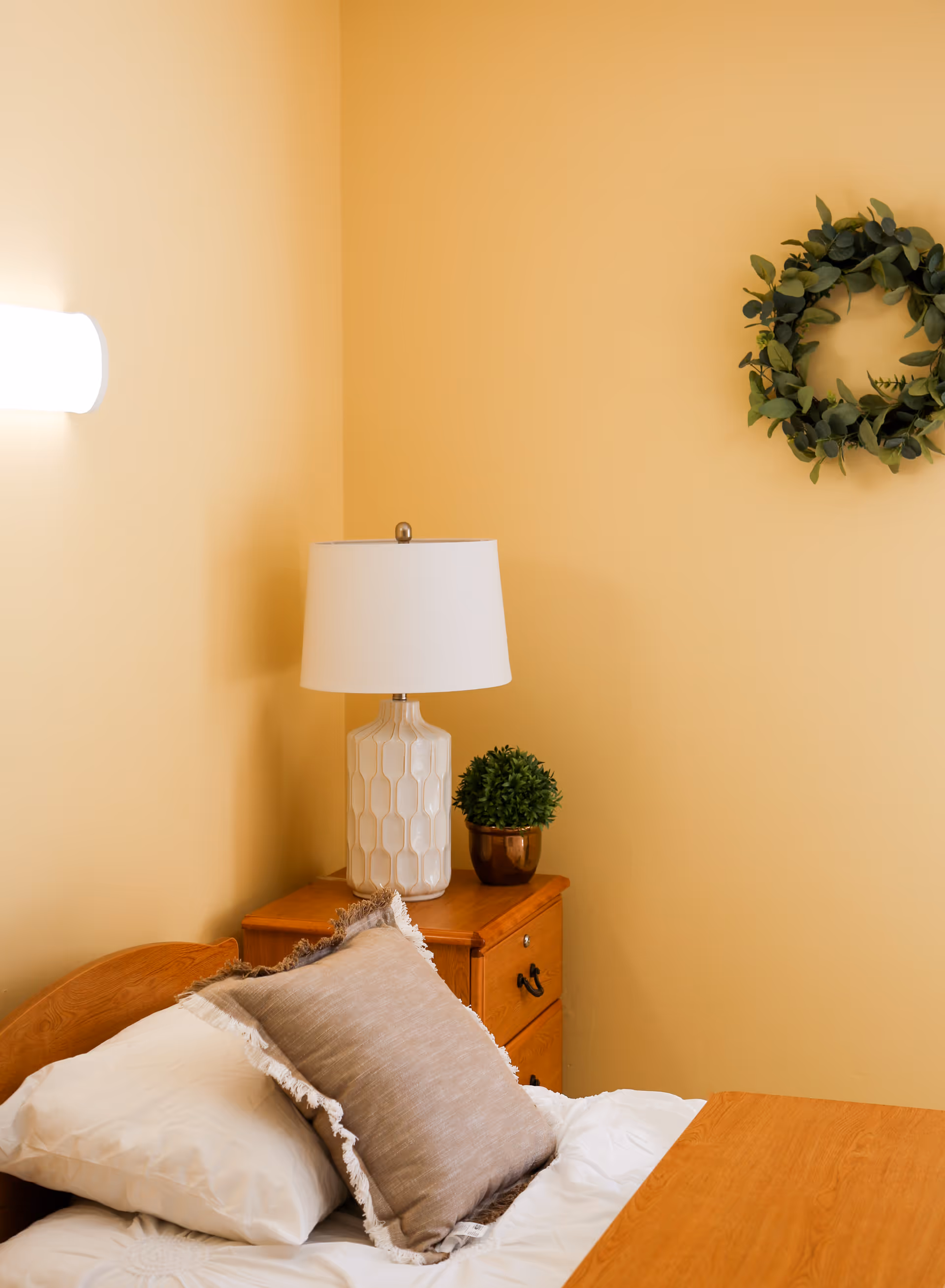 Bedroom corner with a bed and pillow, wooden nightstand holding a lamp and small potted plant, and a green wreath on a yellow wall.