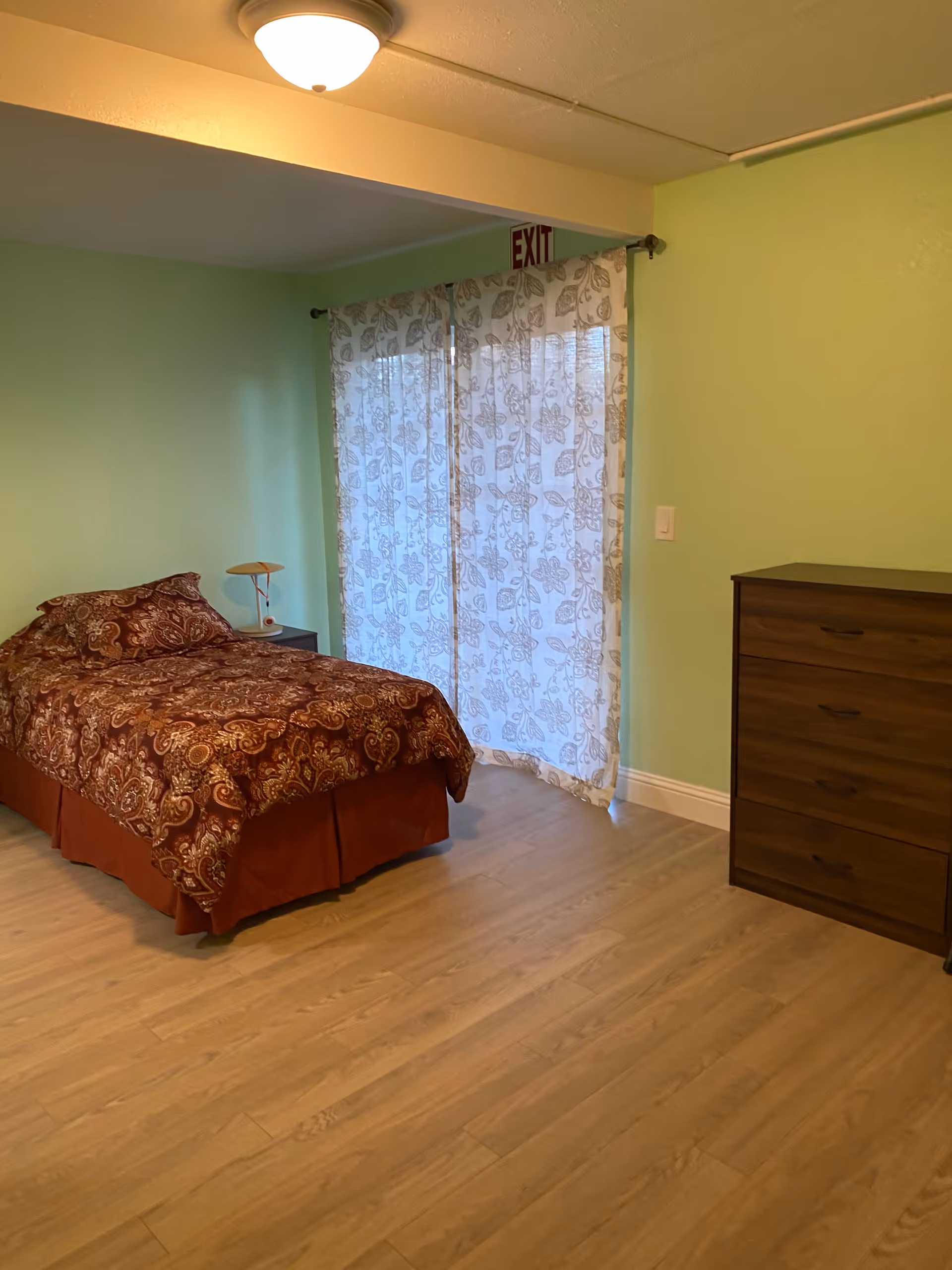 A bedroom with light green walls and wood flooring featuring a single bed with a patterned brown and red bedspread, a small nightstand with a lamp, a wooden dresser, and sheer floral curtains covering a window or door with an exit sign above.