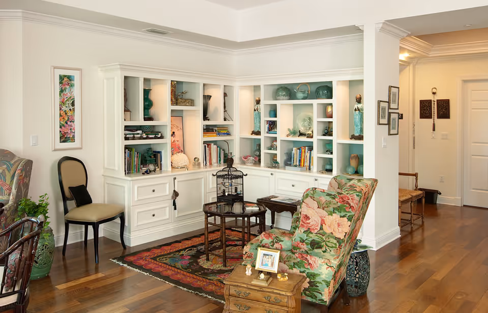 A cozy living room area with wooden flooring, a floral upholstered armchair, a small wooden side table with framed photos and decorative items, a glass-top coffee table with a birdcage, and built-in white shelves filled with books, vases, and decorative objects. The walls are light-colored, and there is a hallway visible in the background.